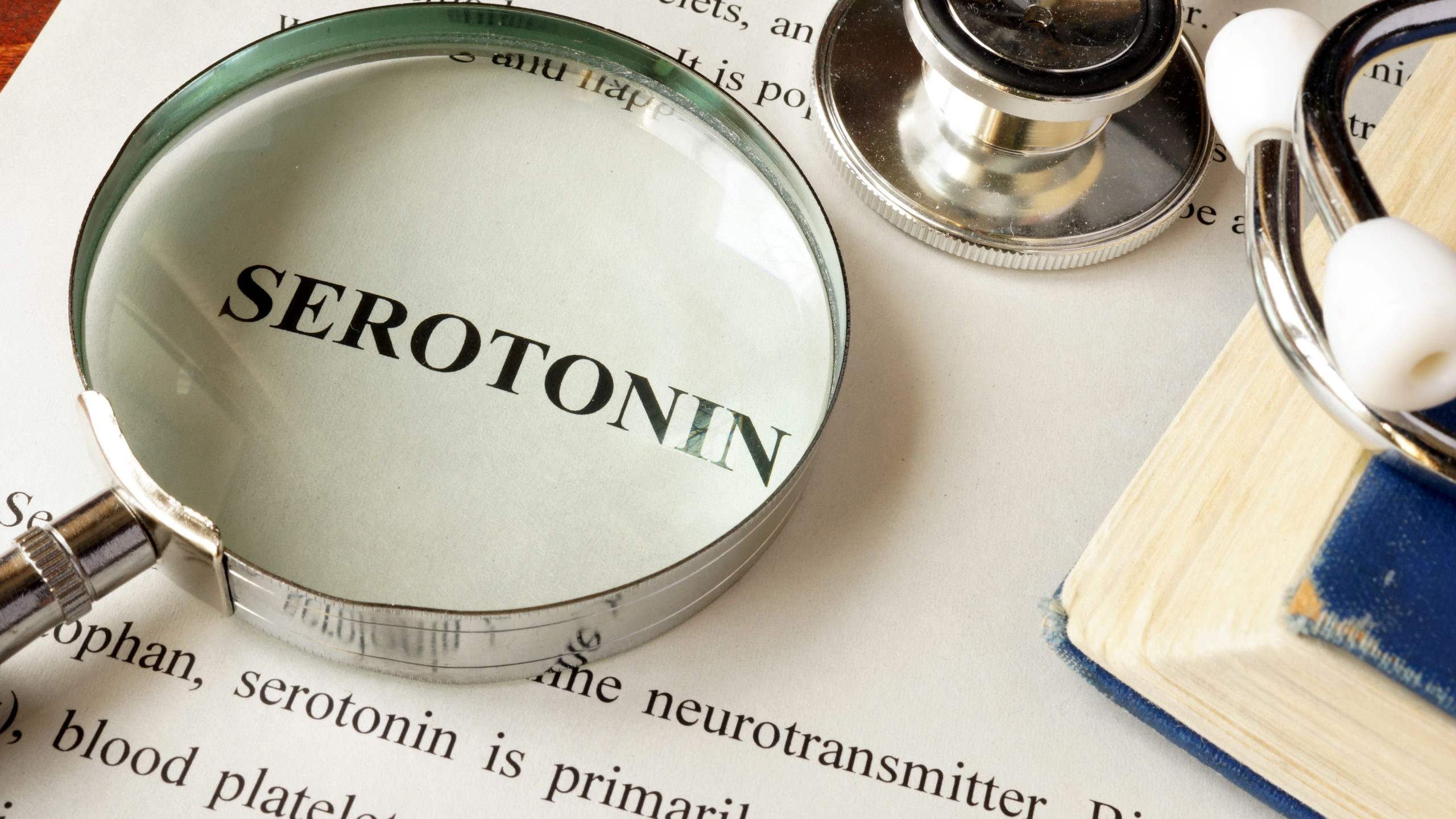 A magnifying glass positioned over a book with the word 'serotonin' highlighted, illustrating a focus on the study or understanding of serotonin.